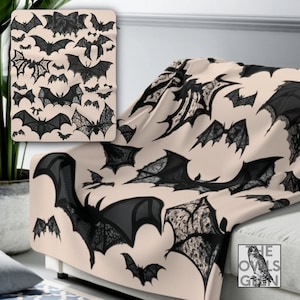 May include: A soft, beige fleece throw with a repeating pattern of black lace bats. The throw is folded on a sofa with a cushion and a small sign that reads "The Owls Gettin".