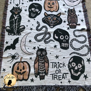 Puede incluir: Una manta de tejido blanco con un tema de Halloween negro y naranja. La manta presenta im&aacute;genes de gatos, b&uacute;hos, calabazas, calaveras, serpientes, murci&eacute;lagos y estrellas. El texto "Trick or Treat" est&aacute; tejido en la manta.