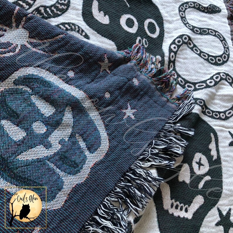 Puede incluir: Un tapiz tejido en blanco y negro con un dise&ntilde;o de calavera y serpiente. El tapiz tiene un borde de flecos y presenta un peque&ntilde;o logotipo de b&uacute;ho en la esquina inferior izquierda.