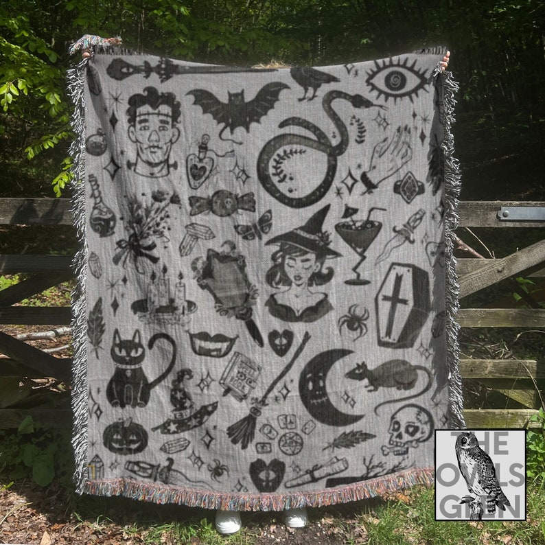 May include: A gray woven throw blanket with a black and white pattern of various spooky and witchy imagery, including a cat, a snake, a skull, a witch, a coffin, a crescent moon, a broom, a potion bottle, a spider, a bat, a hand, a dagger, a feather, a candle, a book, a heart, a pumpkin, a crystal, a mirror, a dice, a feather, and a snake. The blanket has fringe on all sides. The text "THE OWLS GLOW" is visible in the bottom right corner of the image.
