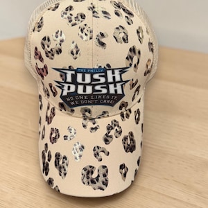 May include: A beige trucker hat featuring a brown and black leopard print pattern. The front panel displays a graphic with the text THE PHILLY TUSH PUSH NO ONE LIKES IT WE DON'T CARE in blue, white, and black lettering on a shield-shaped background.