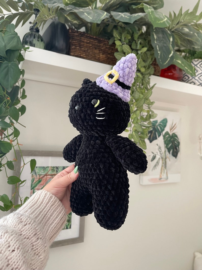 May include: Black crocheted plush cat wearing a purple witch hat with a yellow buckle. The cat has black eyes and a white stitched nose and whiskers.