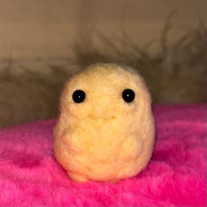 May include: A small, round, pale yellow needle-felted creature with a soft, fuzzy texture. It features two small, round, black bead eyes positioned symmetrically on its face and two tiny, rounded nubs serving as arms on its front side.