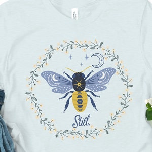 May include: A light blue t-shirt with a floral wreath design featuring a stylized bee with a moon and stars. The text "Still" is printed below the bee.