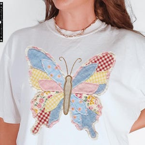 Peut inclure: T-shirt blanc avec un motif de papillon patchwork color&eacute;. Le papillon pr&eacute;sente des tissus bleus, roses, jaunes et &agrave; motifs floraux. Le t-shirt porte les mentions "COMFORT COLORS" et "WHITE". Le logo de la marque "LL&G" est en haut &agrave; gauche.