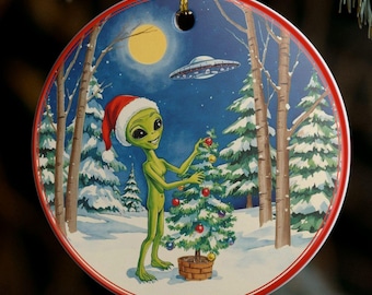 Puede incluir: Adorno navideño redondo con un extraterrestre verde con gorro de Papá Noel, decorando un árbol de Navidad en un bosque nevado. Un OVNI sobrevuela el cielo nocturno con luna llena. El adorno tiene un borde rojo y está colgado de un hilo dorado.
