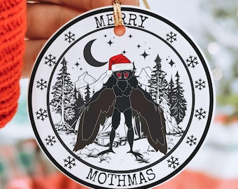May include: A white round Christmas ornament with a black and white illustration of a mothman wearing a Santa hat. The ornament says "Merry Mothmas" in black text.