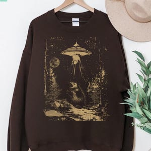 May include: A dark brown sweatshirt with a gold-toned graphic of a UFO shining a light on a creature in a forest scene. The sweatshirt has long sleeves and a crew neck. The text "LOVE LIGHT GRACE" is visible in the upper left corner.