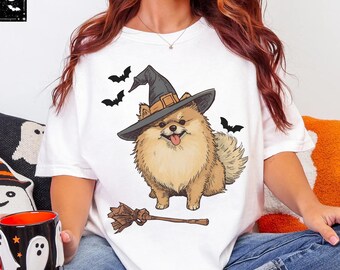 May include: White t-shirt featuring a cartoon Pomeranian dog wearing a witch hat and surrounded by bats. A broom lies below the dog. The shirt is part of a Halloween-themed collection.