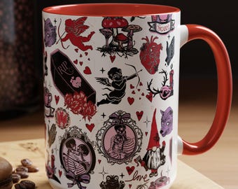 May include: A white ceramic mug with a red interior and handle, decorated with gothic-themed illustrations. The design includes cherubs, hearts, mushrooms, skeletons, and the word "love". A spoon and a macaron are in the foreground.