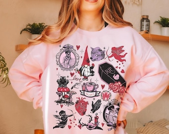 Può includere: Una felpa rosa chiaro con illustrazioni a tema gotico. Il design include uno gnomo, un lupo, un cupido, una bara e altri elementi. Le parole "Love Light and Grace" sono stampate in basso a sinistra.