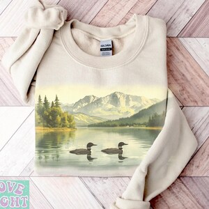 May include: Beige sweatshirt featuring a scenic lake view with two loons swimming. The landscape includes mountains, trees, and a calm lake. The sweatshirt has a crew neck and long sleeves. The text "LOVE LIGHT GRACE" is visible in the lower left corner.
