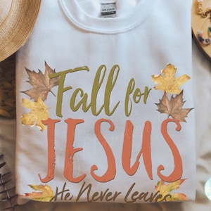 May include: White sweatshirt with the text "Fall for JESUS He Never Leaves" in olive green and orange, decorated with autumn leaves. The sweatshirt is a Gildan brand.