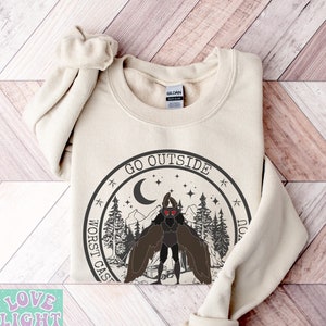 May include: A beige sweatshirt featuring a graphic print of a creature with red eyes and wings standing in a forest with a crescent moon and stars. The text "GO OUTSIDE WORST CASE YOU" is printed in a circle around the creature.