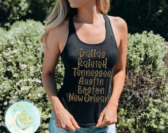 May include: A black tank top with a gold glitter design featuring the names of cities: Dallas, Raleigh, Tennessee, Austin, Boston, and New Orleans.
