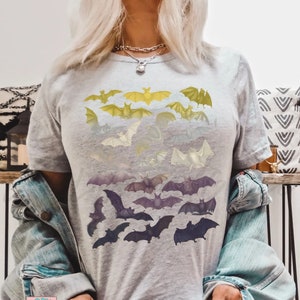 May include: A gray t-shirt with a graphic of bats in a gradient pattern of yellow, white, and purple. The bats are arranged in rows, with the largest bats at the bottom and the smallest bats at the top. The shirt is being worn by a person with long blonde hair.