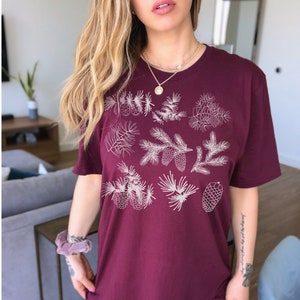 May include: A maroon t-shirt with a white design of pine branches and cones. The shirt is oversized and has a relaxed fit.