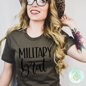 May include: A woman wearing a green t-shirt with the text "MILITARY brat" in black lettering. She is wearing a leopard print hat and glasses.