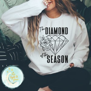May include: White sweatshirt with black text and graphic. The text reads "The Diamond of the Season" and features a diamond shape with a floral design.