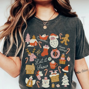 May include: A gray t-shirt with a Christmas-themed design featuring a variety of festive icons, including a gingerbread man, a Santa Claus, a reindeer, a Christmas tree, and a stocking. The text "Let it Snow" is printed on the shirt. The shirt is being worn by a person with a gold necklace and blue jeans.