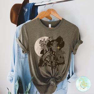May include: An olive green t-shirt featuring a detailed graphic of a wolf howling at the moon, surrounded by floral and celestial elements. The design is in shades of brown and tan, creating a nature-inspired aesthetic. The shirt is hanging on a wooden hanger.