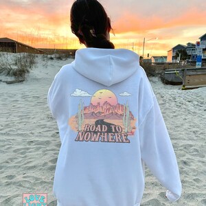 May include: White hoodie featuring a graphic print of a desert landscape with a road and the text "Road to Nowhere". The graphic is in pink, orange, and yellow colours.