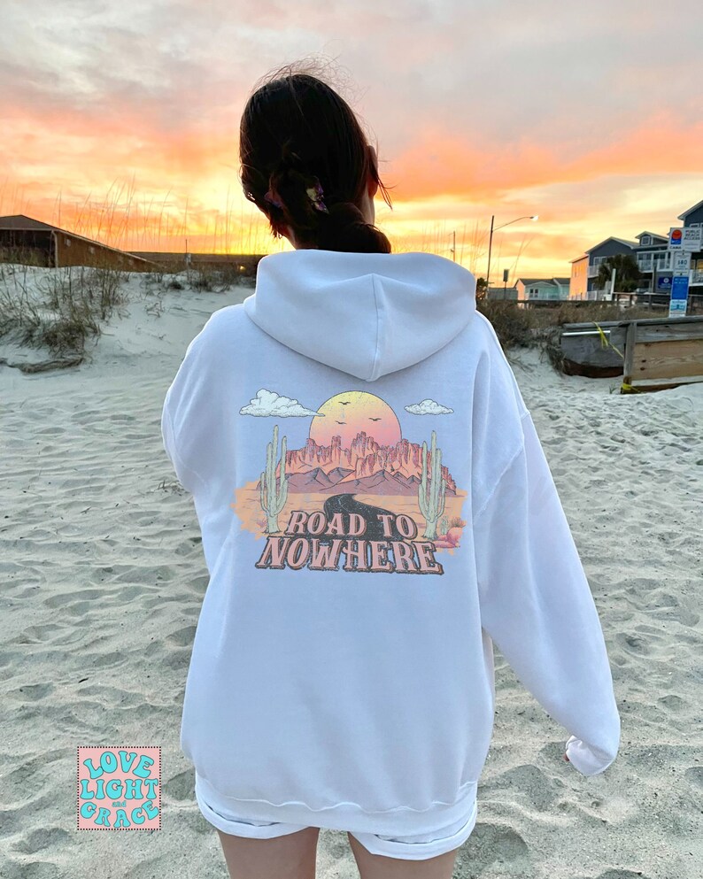 May include: White hoodie featuring a graphic print of a desert landscape with a road and the text "Road to Nowhere". The graphic is in pink, orange, and yellow colours.