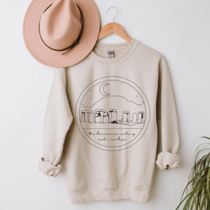 May include: A beige sweatshirt with a black and white illustration of Stonehenge, a crescent moon, and stars. The text "the stones are calling and i must go" is printed below the illustration.