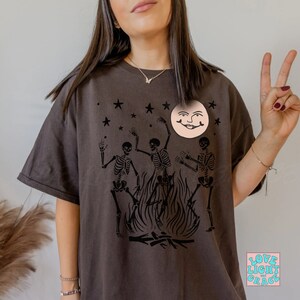 May include: A dark brown t-shirt with a graphic print of three skeletons dancing around a bonfire with a smiling moon face above them. The text "LOVE LIGHT GRACE" is printed in a square on the bottom right corner of the shirt.