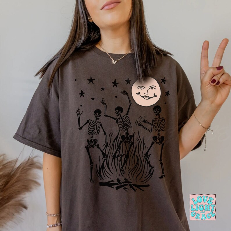 May include: A dark brown t-shirt with a graphic print of three skeletons dancing around a bonfire with a smiling moon face above them. The text "LOVE LIGHT GRACE" is printed in a square on the bottom right corner of the shirt.
