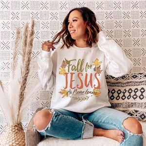 May include: White long-sleeve sweatshirt with the text "Fall for JESUS He Never Leaves" in orange and green, surrounded by fall leaves. The sweatshirt is a comfortable, casual style.