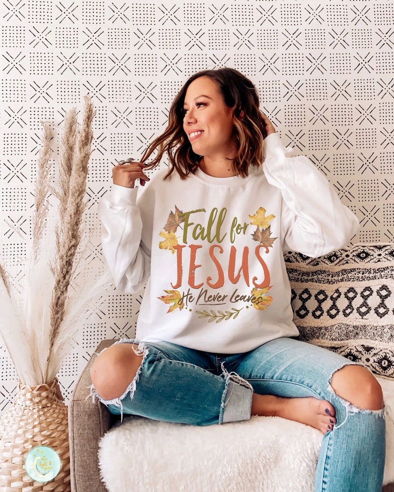 May include: White long-sleeve sweatshirt with the text "Fall for JESUS He Never Leaves" in orange and green, surrounded by fall leaves. The sweatshirt is a comfortable, casual style.