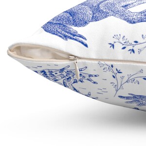 May include: White throw pillow with a blue illustration of a deer and floral elements. The pillow has a zipper closure and is made of a textured fabric. The design is repeated across the pillow's surface.