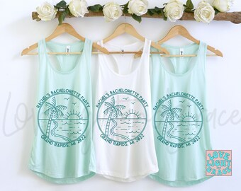 May include: Three light blue tank tops with a teal green graphic of a palm tree, sun, and ocean waves. The text "Rachel's Bachelorette Party Grand Rapids, MI 2022" is printed on each tank top.