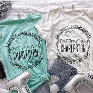 May include: Two white t-shirts with black text and a graphic of a city skyline with a crescent moon and stars. The text on the shirts reads "Brittany's Bachelorette Charleston SC Last Fling Down King".