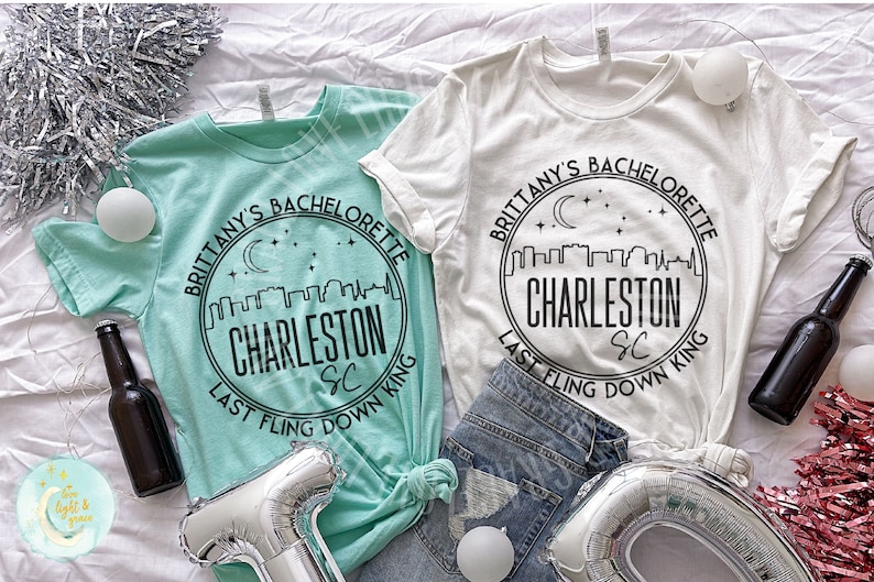 May include: Two white t-shirts with black text and a graphic of a city skyline with a crescent moon and stars. The text on the shirts reads "Brittany's Bachelorette Charleston SC Last Fling Down King".