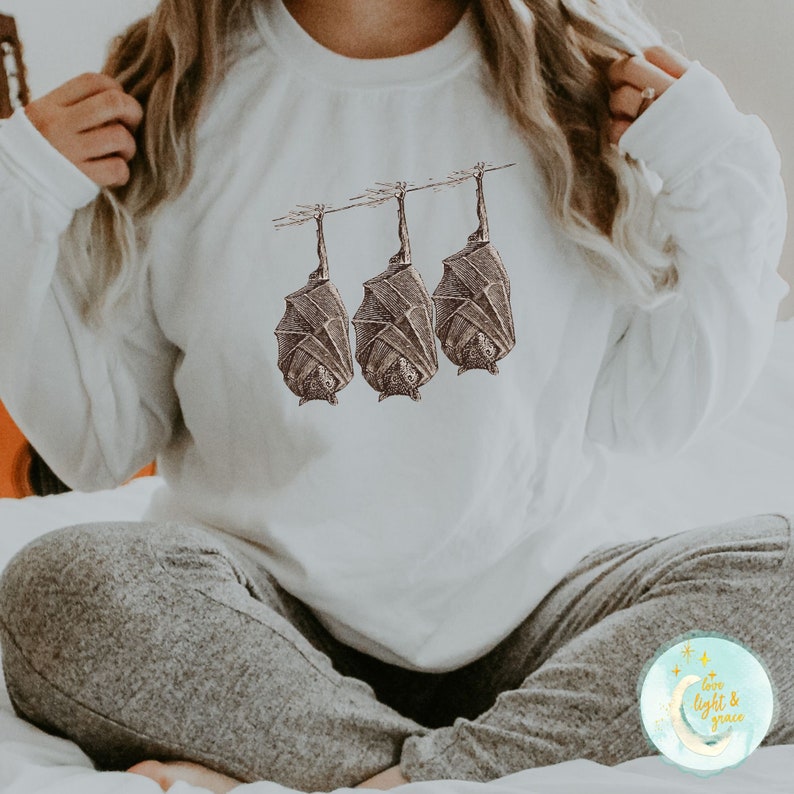 May include: White sweatshirt with a graphic of three bats hanging upside down. The bats are drawn in a vintage style with brown and black details. The sweatshirt has a crew neck and long sleeves.