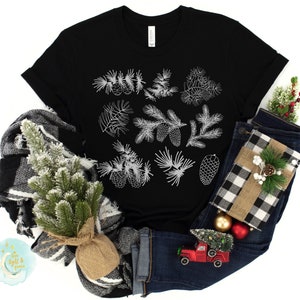 May include: Black t-shirt with a white design of pine branches and cones. The shirt is displayed on a white background with a small Christmas tree, a plaid scarf, and blue jeans.