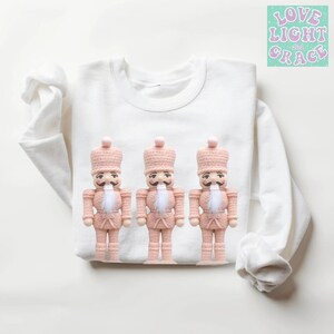 May include: A white sweatshirt with three pink nutcracker figures. The nutcrackers have black mustaches and white beards. The text "LOVE LIGHT and GRACE" is in the upper right corner. The sweatshirt is made of a soft material.