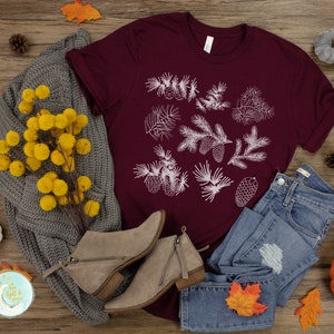 May include: A maroon t-shirt with a white design of pine branches and cones. The shirt is laid out on a wooden surface with a gray sweater, blue jeans, fall leaves, and a small pumpkin.