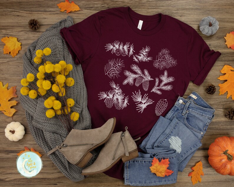 May include: A maroon t-shirt with a white design of pine branches and cones. The shirt is laid out on a wooden surface with a gray sweater, blue jeans, fall leaves, and a small pumpkin.