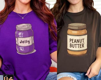 May include: Two t-shirts, one purple and one brown, each with a food-themed graphic. The purple shirt features a jar of jelly, while the brown shirt displays a jar of peanut butter. Both shirts have short sleeves.