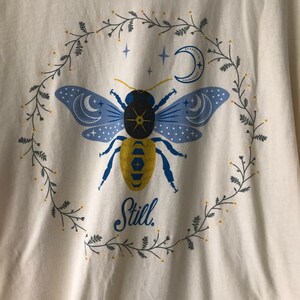 May include: A white t-shirt with a blue and yellow bee illustration surrounded by a wreath of green leaves and white flowers. The bee has a black body and blue wings with crescent moons and stars. The text "Still." is printed below the bee.