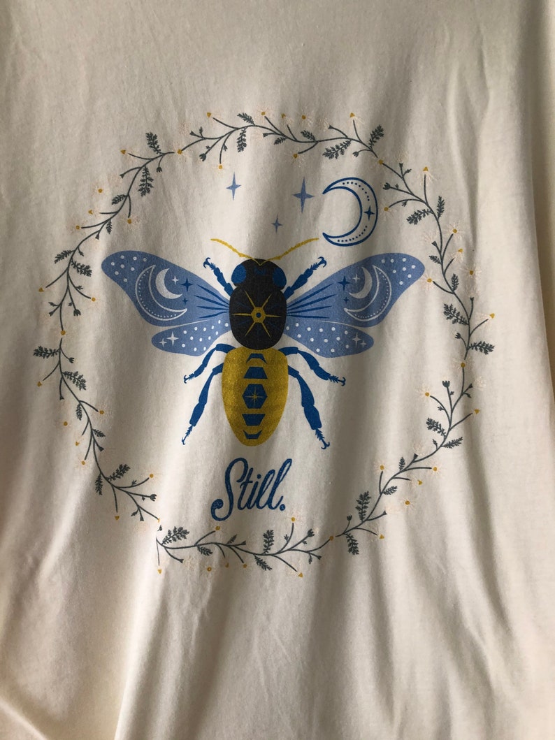 May include: A white t-shirt with a blue and yellow bee illustration surrounded by a wreath of green leaves and white flowers. The bee has a black body and blue wings with crescent moons and stars. The text "Still." is printed below the bee.
