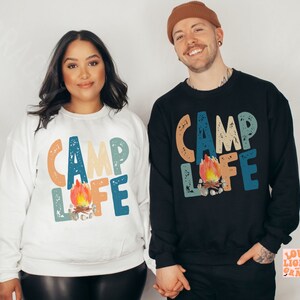May include: Two sweatshirts, one white and one black, each with the words "CAMP LIFE" in colorful, distressed lettering. A campfire graphic is centered below the text. The sweatshirts are casual and suitable for camping or outdoor activities.