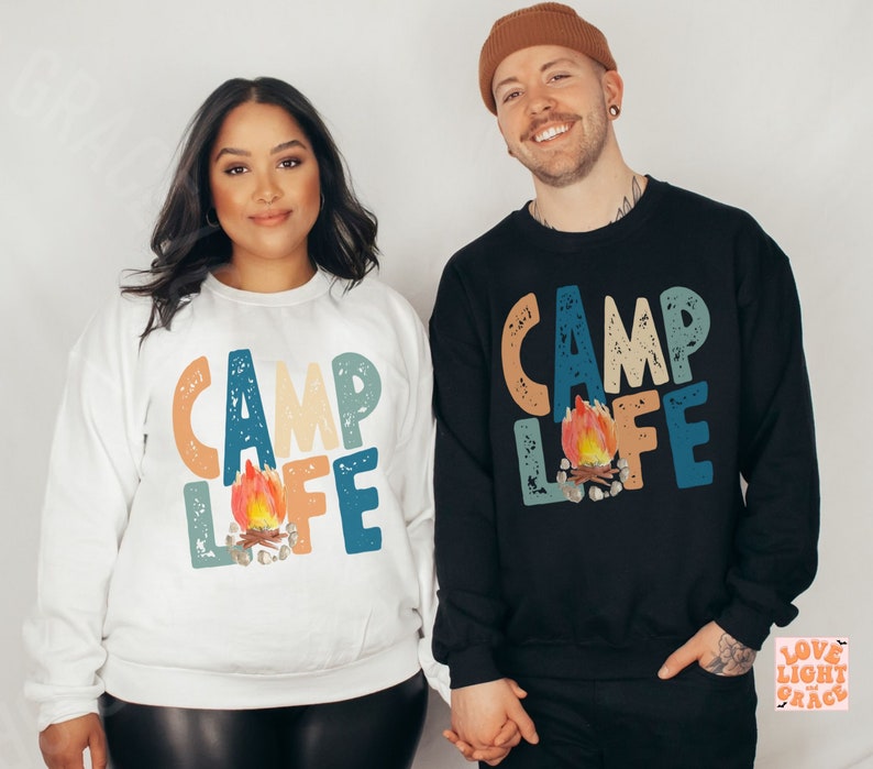 May include: Two sweatshirts, one white and one black, each with the words "CAMP LIFE" in colorful, distressed lettering. A campfire graphic is centered below the text. The sweatshirts are casual and suitable for camping or outdoor activities.