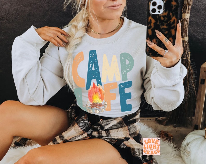 May include: A light gray sweatshirt with the words "CAMP LIFE" in colorful letters and a watercolor campfire graphic. The sweatshirt has a crew neck and long sleeves. The image also includes a phone case and a plaid shirt.
