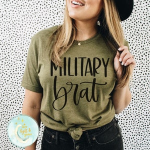 May include: A woman wearing a green t-shirt with the text "MILITARY brat" in black lettering. She is wearing black jeans and a black hat.