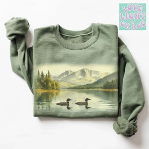 May include: Sage green sweatshirt featuring a scenic lake landscape with two loons. The graphic includes mountains, trees, and water, creating a nature-inspired design. The text "Love Light and Grace" is in the upper right corner.