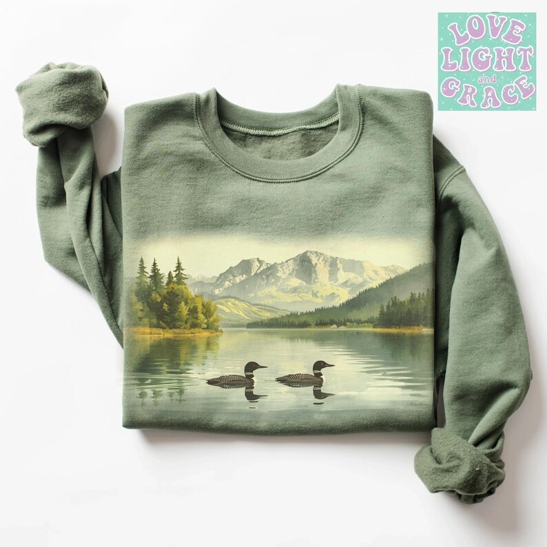 May include: Sage green sweatshirt featuring a scenic lake landscape with two loons. The graphic includes mountains, trees, and water, creating a nature-inspired design. The text "Love Light and Grace" is in the upper right corner.